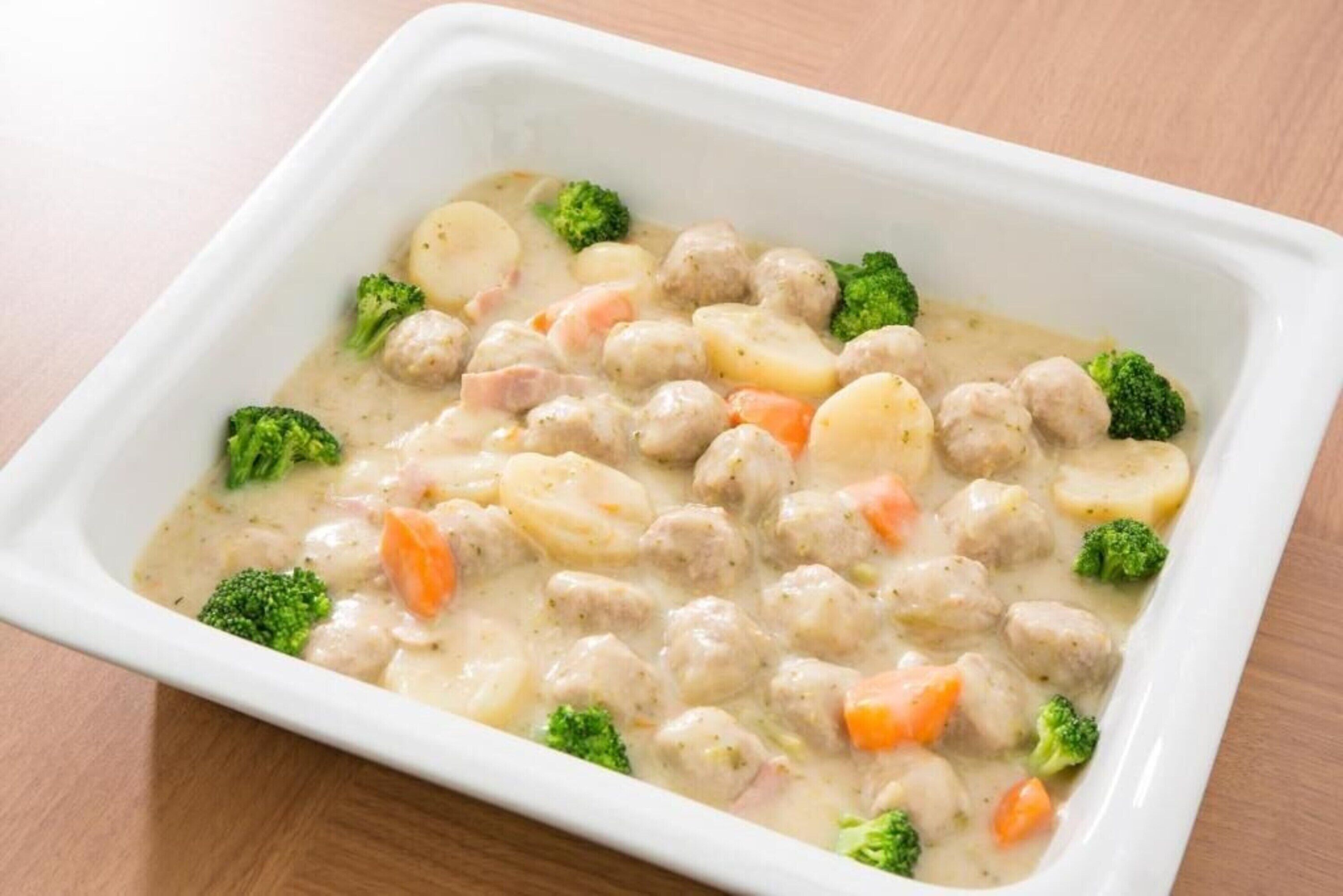 A white rectangular dish containing meatballs, potatoes, carrots, and broccoli in a creamy sauce.