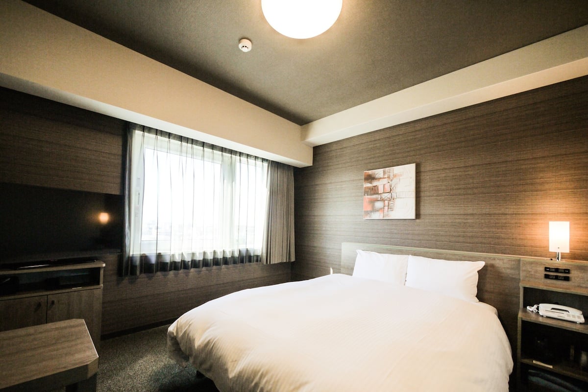 Ein Hotelzimmer mit einem großen Bett, einem Flachbildfernseher, einem Nachttisch, einer Lampe und einem Fenster mit Vorhängen.