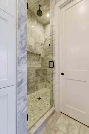 Une douche à l’italienne avec des murs en marbre et une porte en verre, située à côté d’une porte blanche.