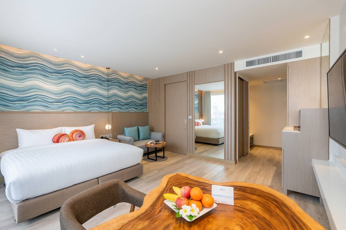 A modern hotel room with a large bed, a wooden dining table with fruit, a sofa, and a TV.