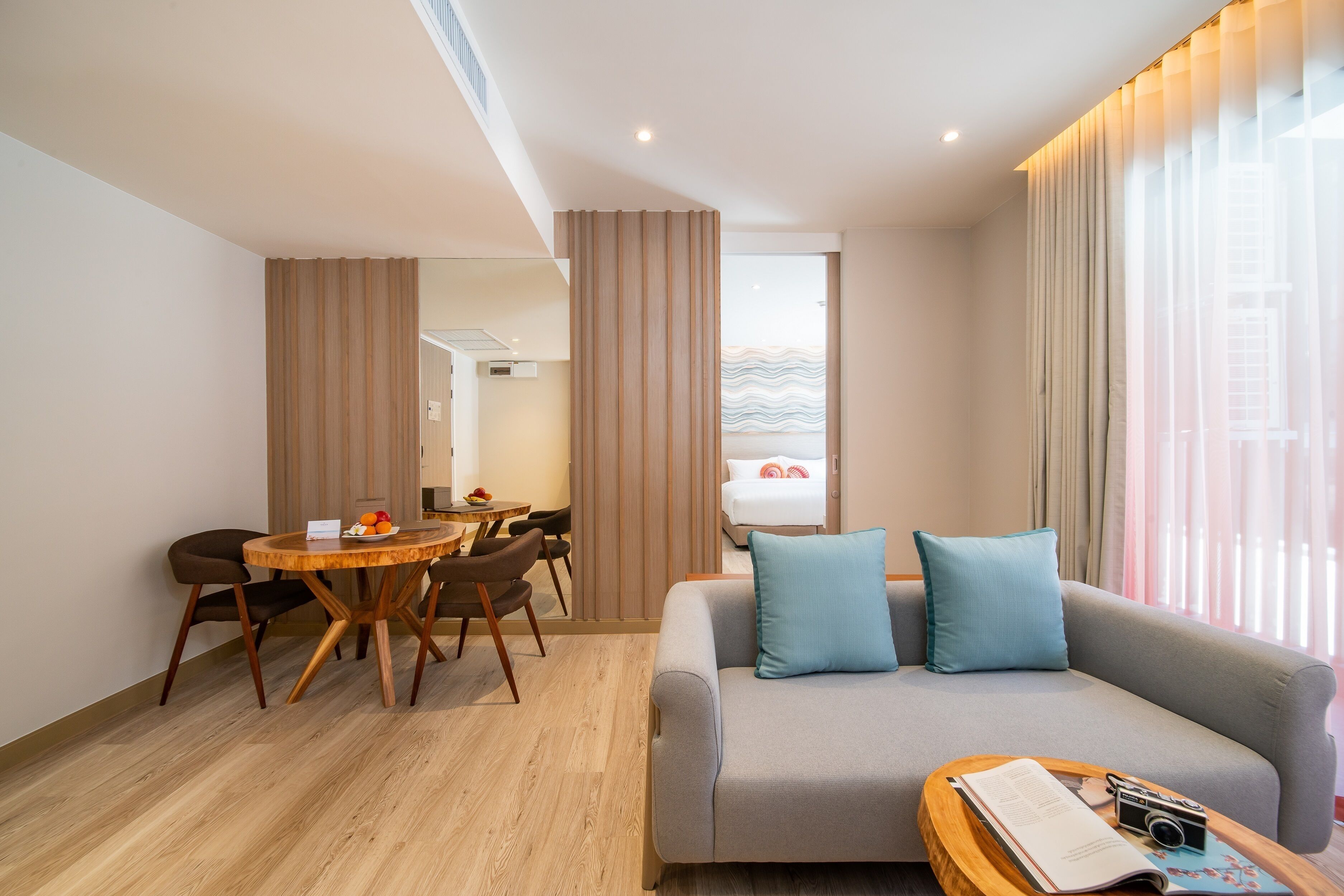A modern hotel room with a grey sofa, a wooden dining table, and a view of the sea through large windows.
