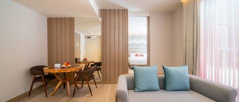 A modern hotel room with a grey sofa, a wooden dining table, and a view of the sea through large windows.