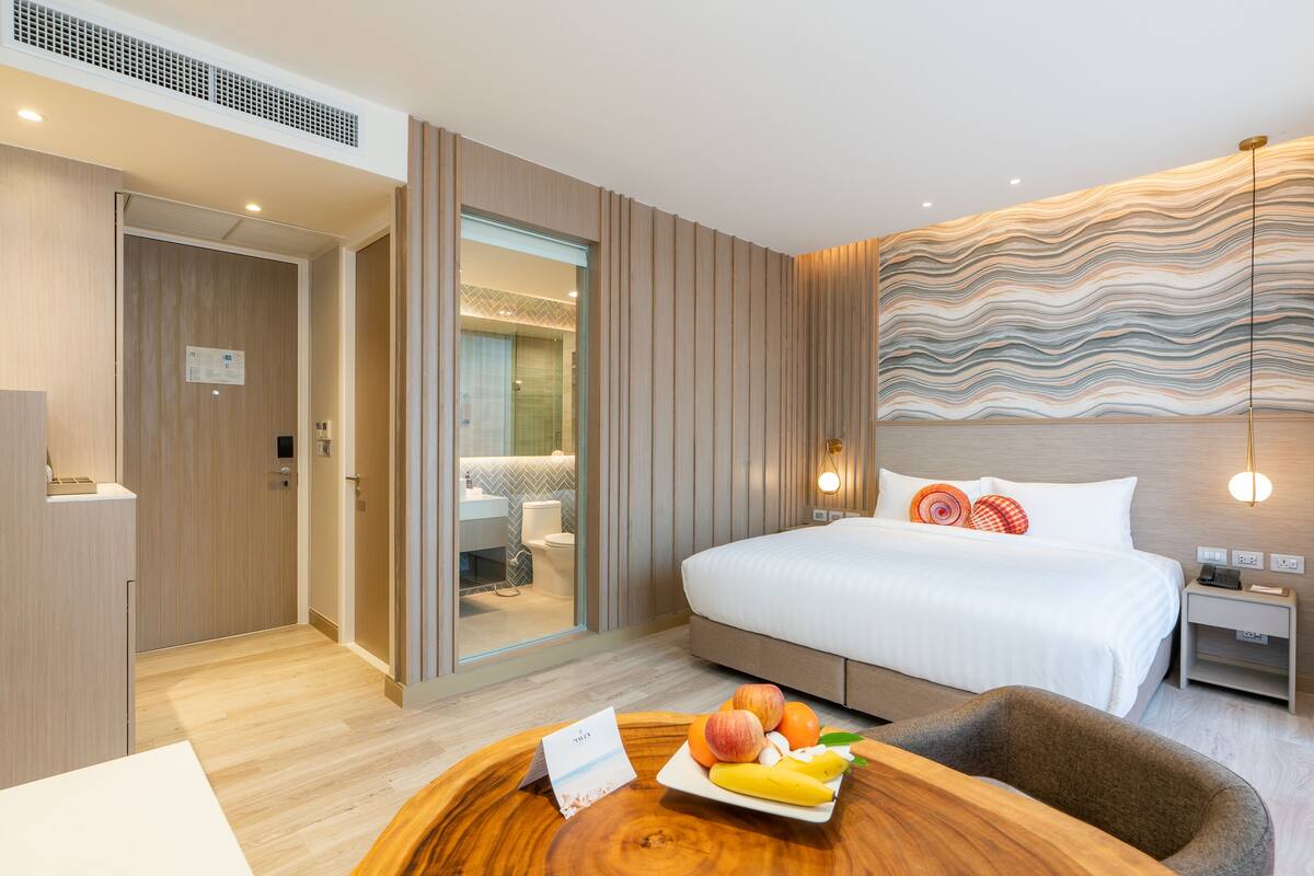A modern hotel room with a wooden floor, a bed with white bedding, a bedside table with a lamp, and a fruit bowl on the side table.