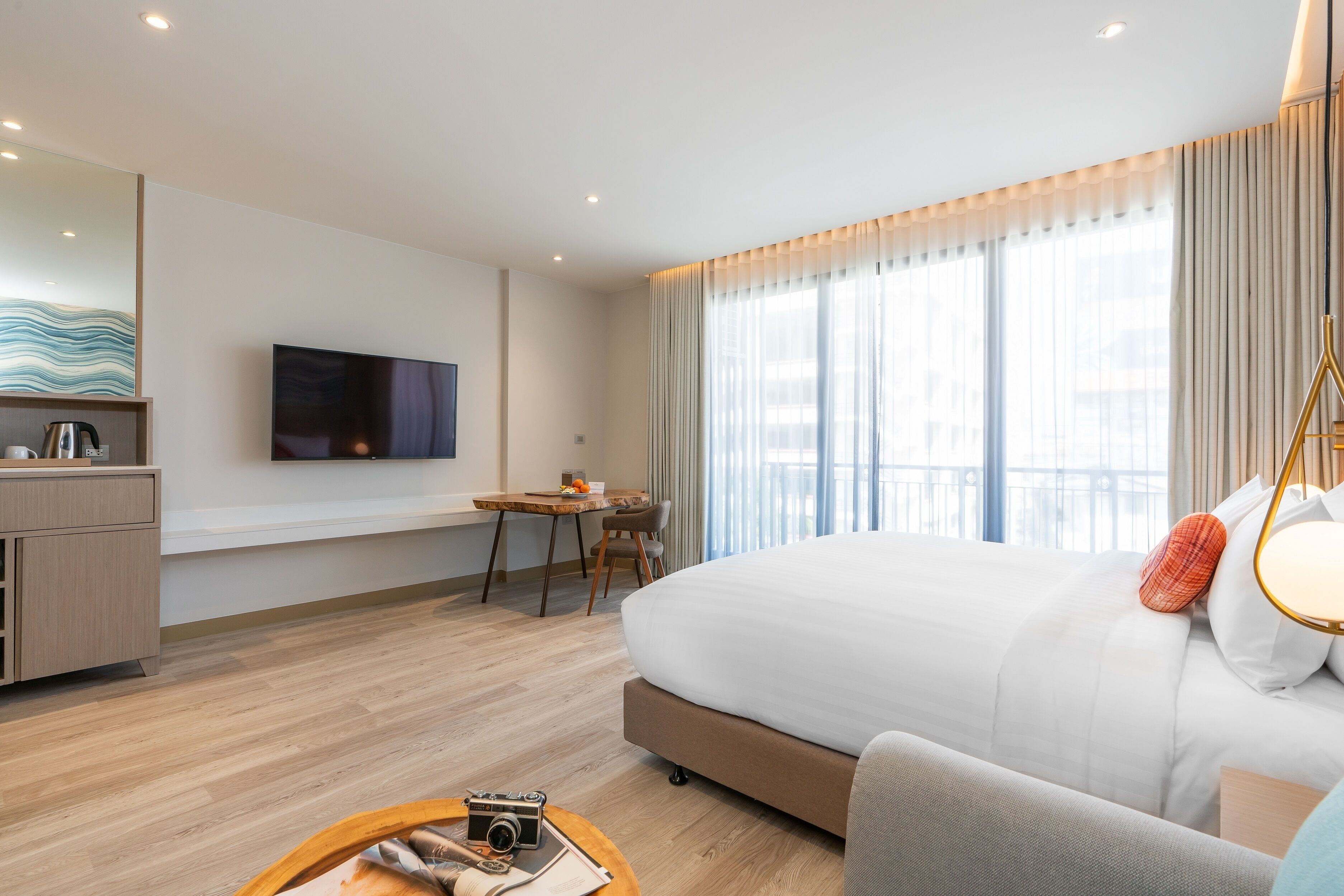 A modern hotel room with a large bed, a desk with a chair, a television mounted on the wall, and a balcony with curtains.