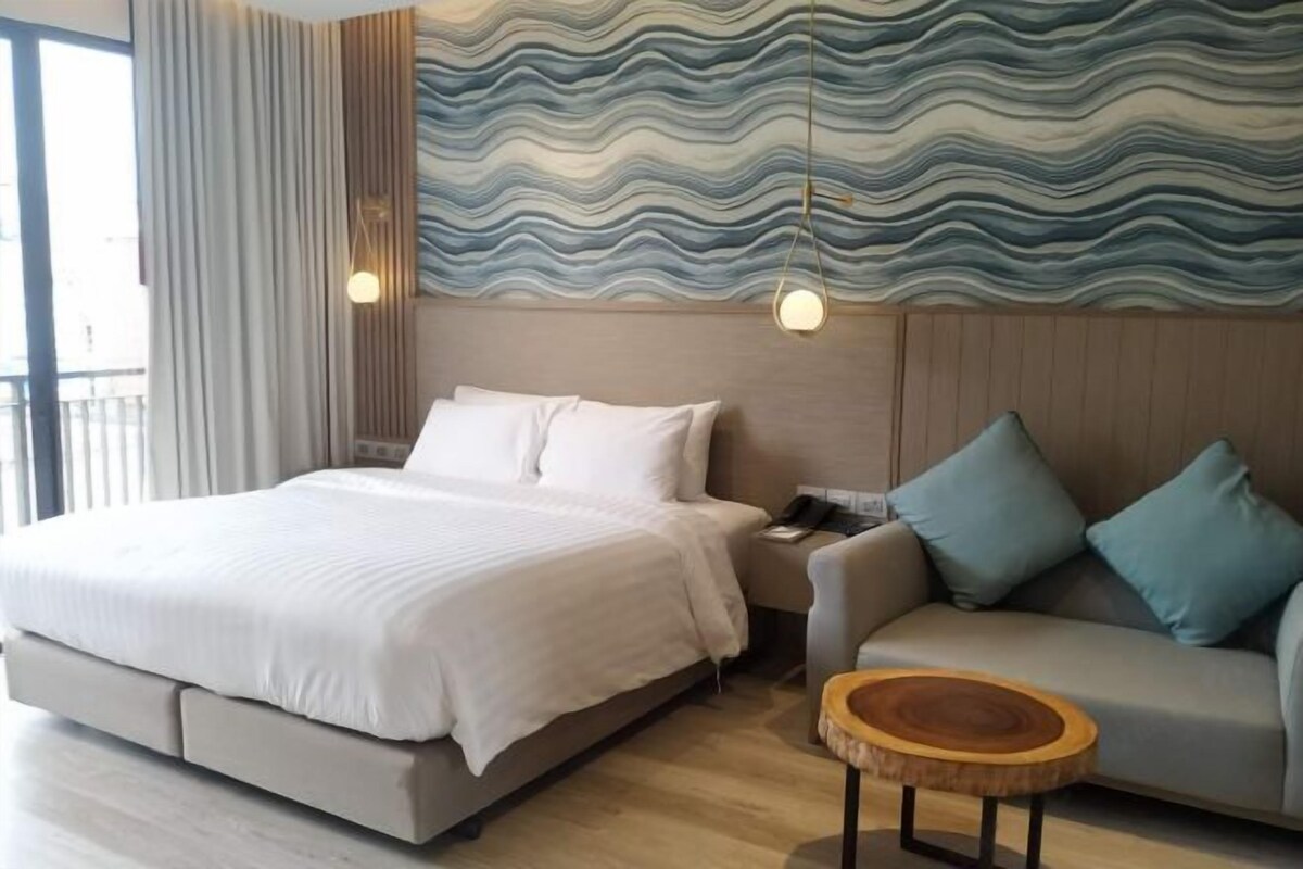 Una habitación de hotel moderna con una cama grande, un sofá, una mesita redonda de madera y un mural texturizado en la pared.