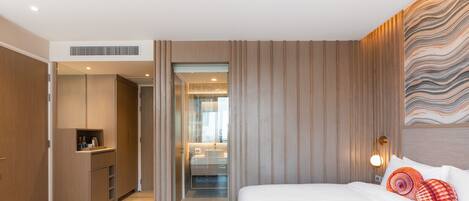 Une chambre d’hôtel moderne avec un plancher en bois, un lit recouvert de draps blancs, une armoire en bois, un bureau et une salle de bain visible à travers une porte en verre.