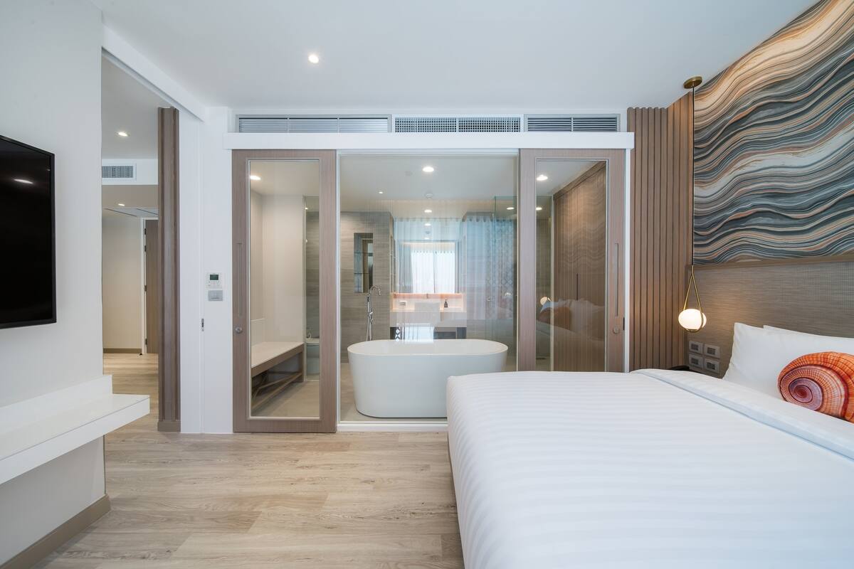 A modern hotel room with a large bed, a bathtub, and a television on the wall.