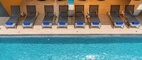 Un área junto a la piscina con tumbonas alineadas, una terraza de madera y una piscina azul cristalina.
