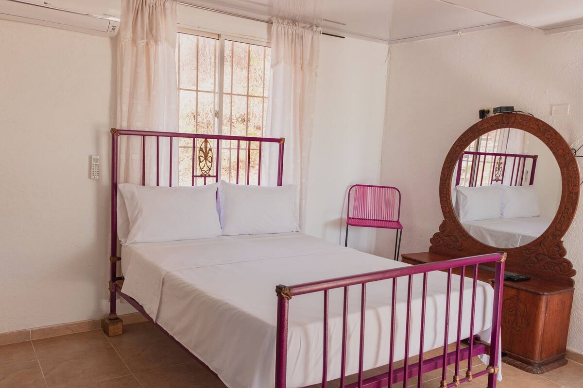 Un dormitorio con cama, una silla, cómoda de madera, espejo y ventana con cortinas.