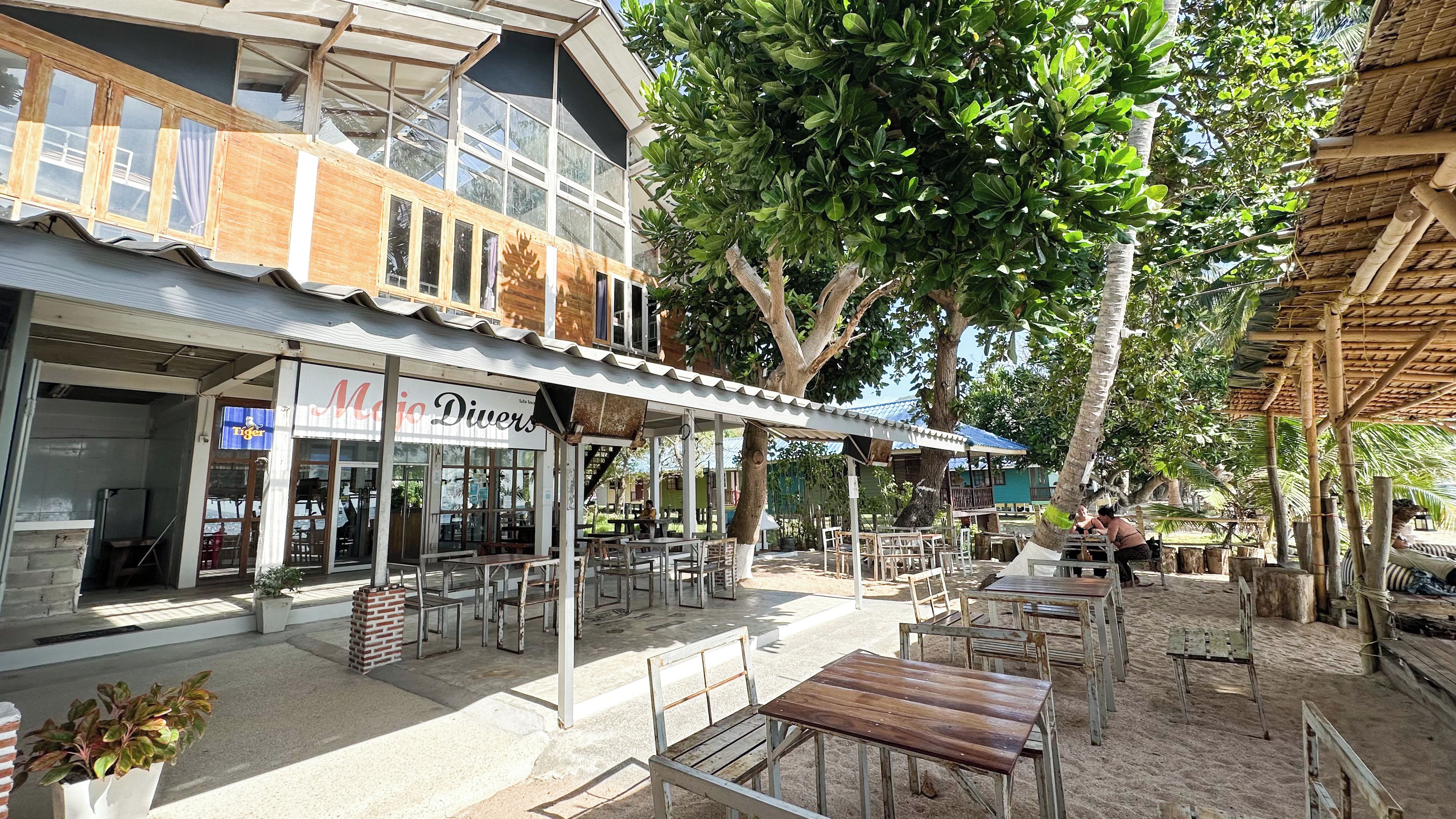 An outdoor cafe with tables and chairs, a building with a sign, and a large tree.
