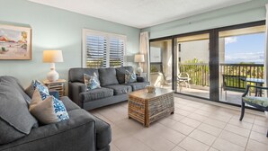 A spacious living room with a grey sofa, a wooden coffee table, and a balcony with a view. - Oceanfront | Pool View | Gorgeously Furnished 3rd Floor Amelia Surf and Racquet Condo (Fernandina Beach)