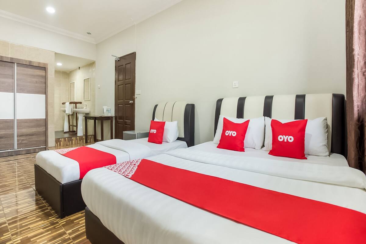 A hotel room with two beds, each with red and white bedding, and a red blanket. There is a wooden wardrobe, a small table with a chair, and a bathroom visible in the background.