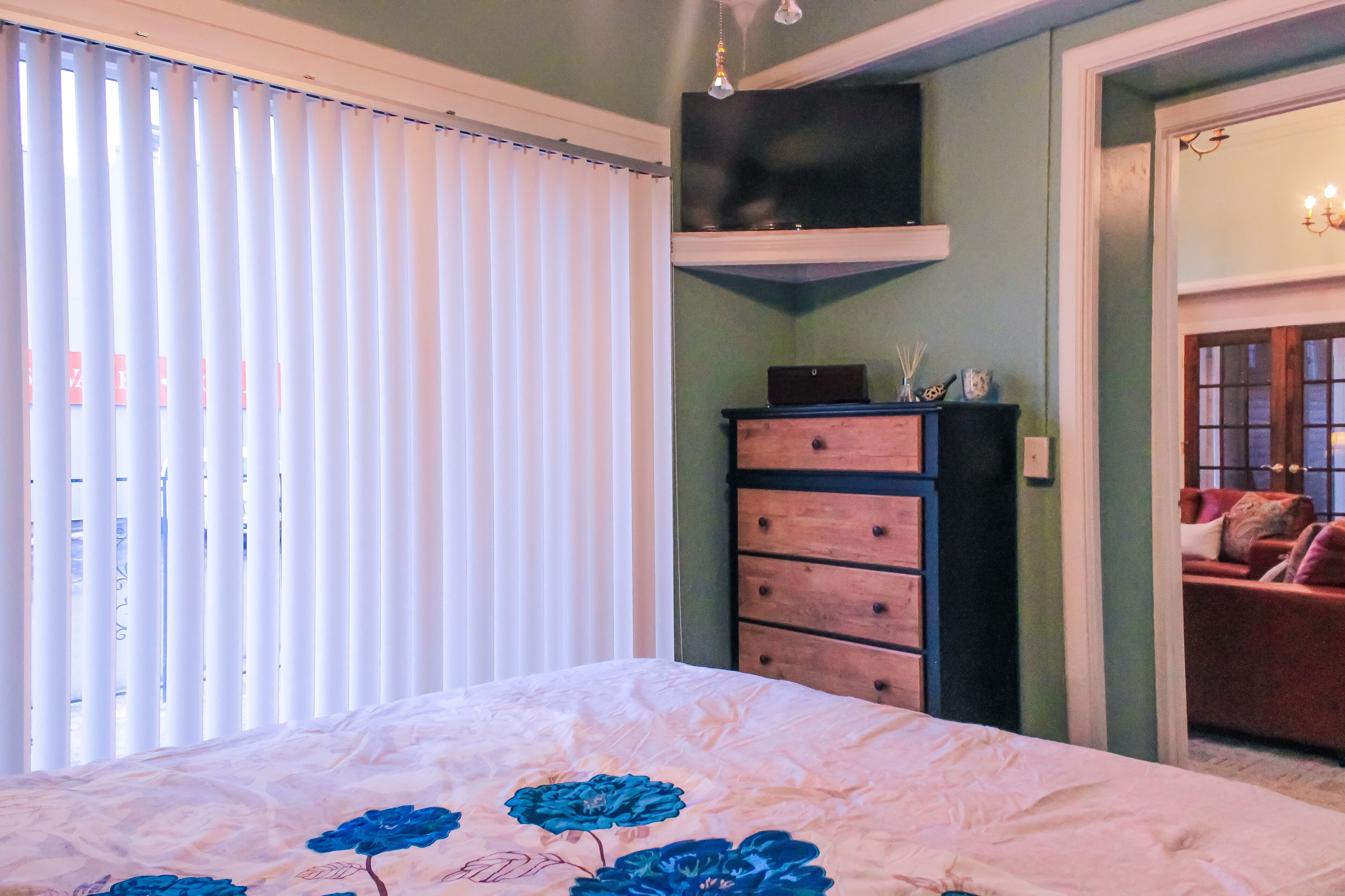 A bedroom with a bed, a dresser, a television on a shelf, and a door leading to another room.