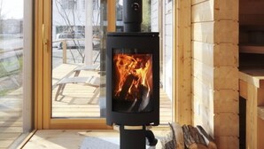 Un intérieur moderne en bois, avec une cheminée centrale, une porte vitrée donnant sur l’extérieur et une pile de bois de chauffage.