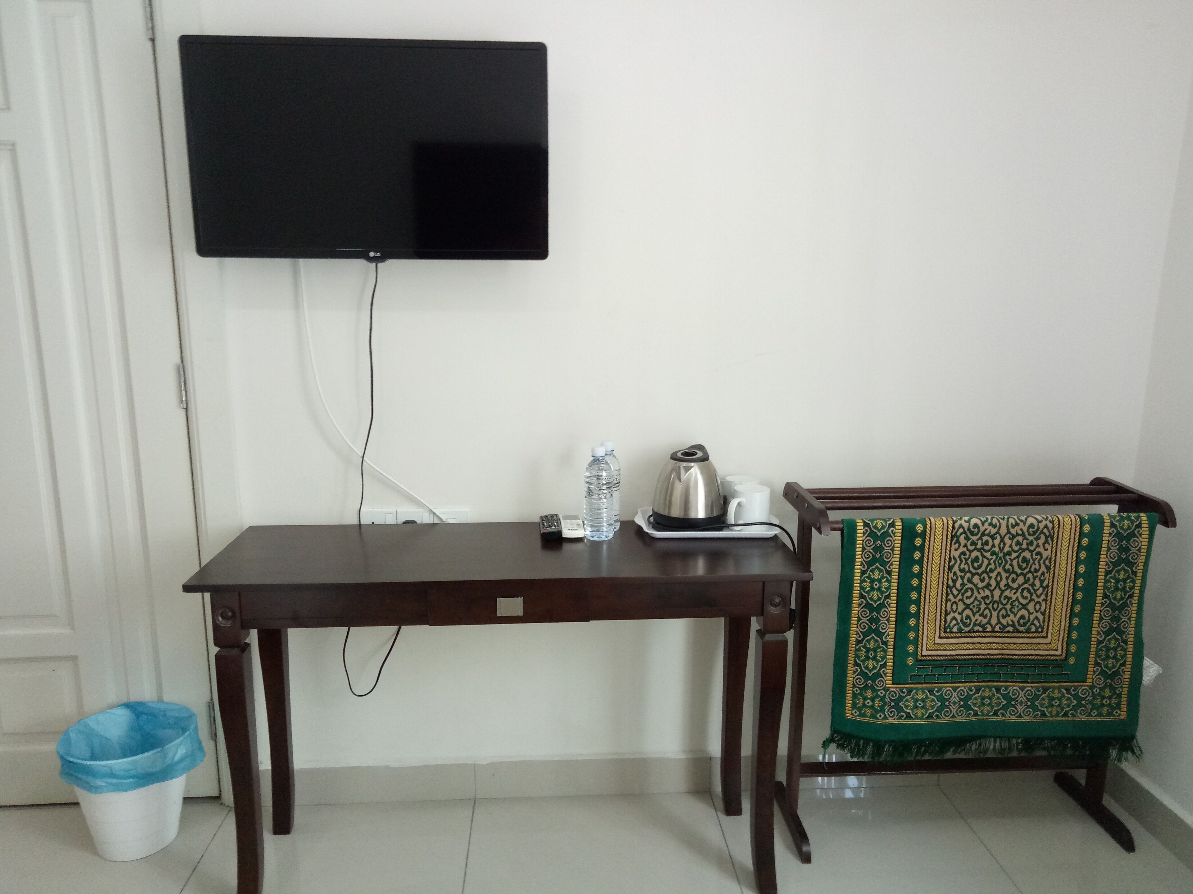 A room with a flat-screen TV mounted on the wall, a wooden table with a kettle, cups, and a bottle, and a patterned chair with a green and gold cover.