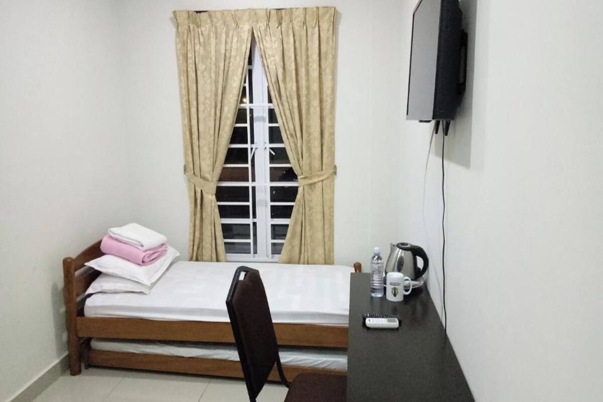 Ein kleines Zimmer mit einem Bett, einem Schreibtisch, einem Sessel, einem Fernseher und einem Fenster mit Vorhängen.