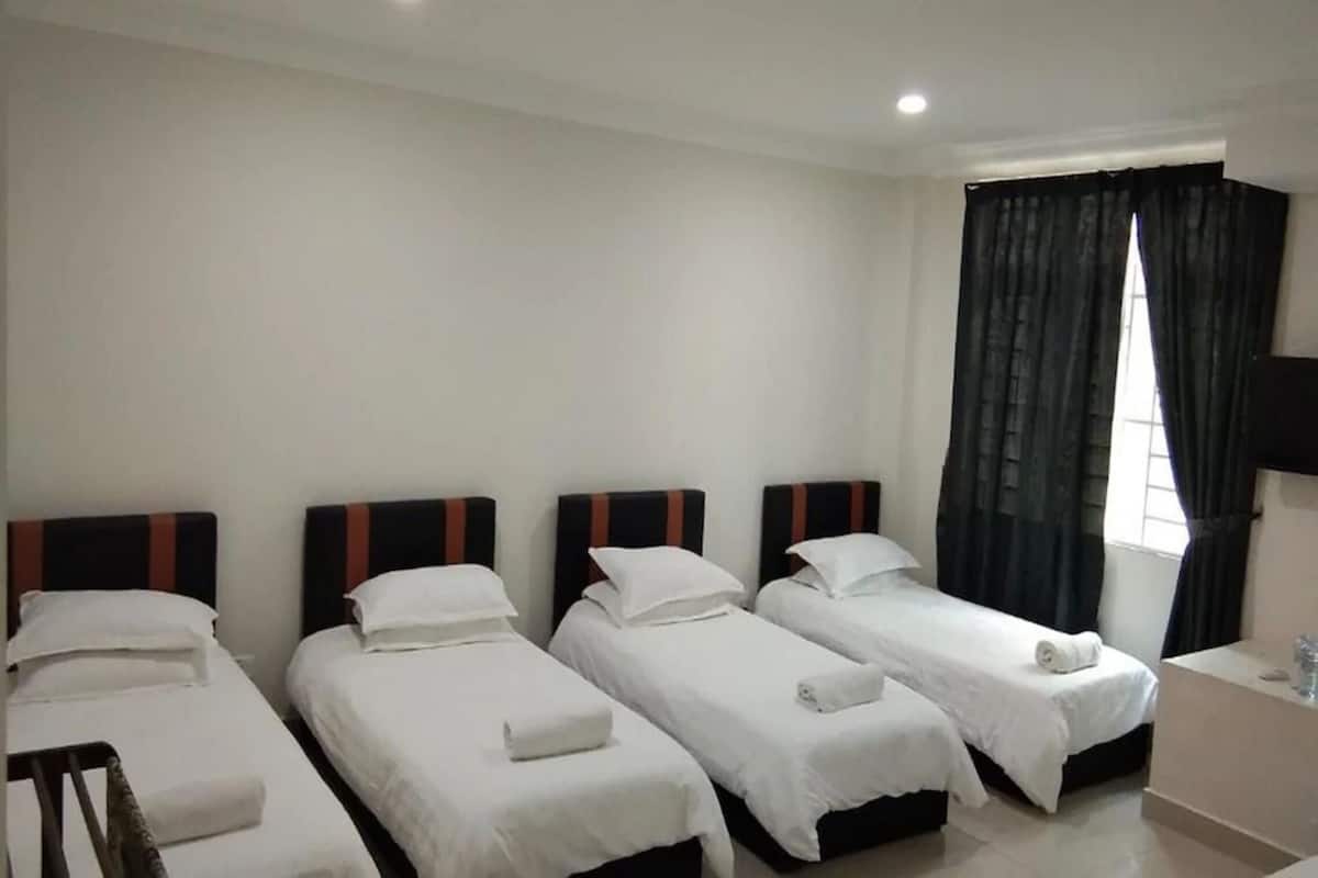 Ein Hotelzimmer mit vier Einzelbetten, jeweils mit weißer Bettdecke und einem gerollten Handtuch. Es gibt ein Fenster mit dunklen Vorhängen, einen Flachbildfernseher und einen kleinen Nachttisch.