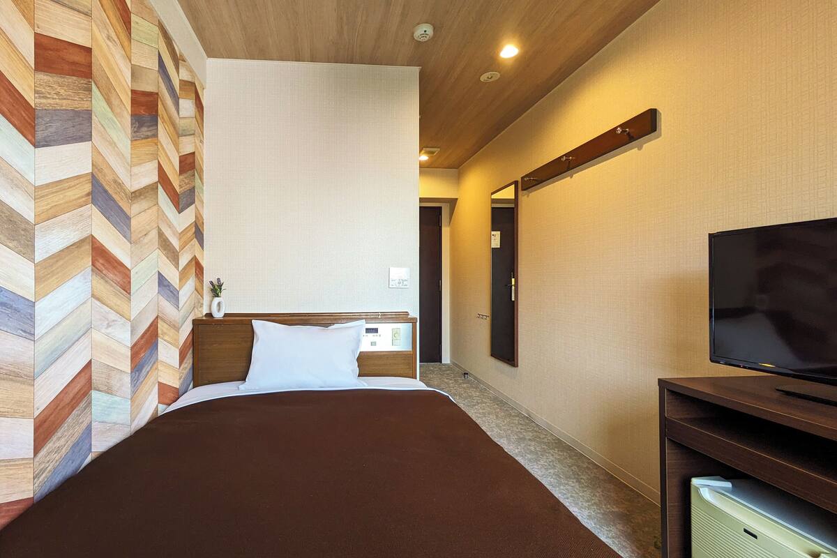 A hotel room with a bed, a TV, a wooden wall panel, a door, and a light fixture.