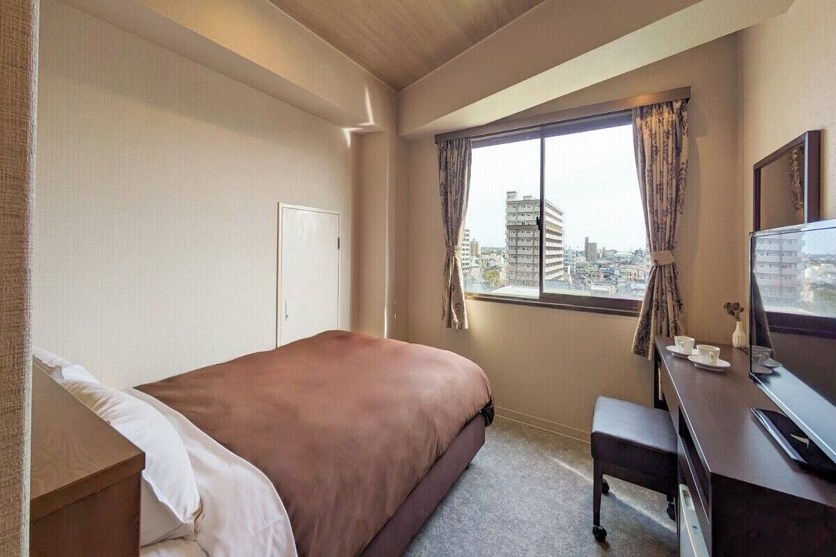 A hotel room with a bed, a desk with a computer, a window with curtains, and a view of buildings outside.