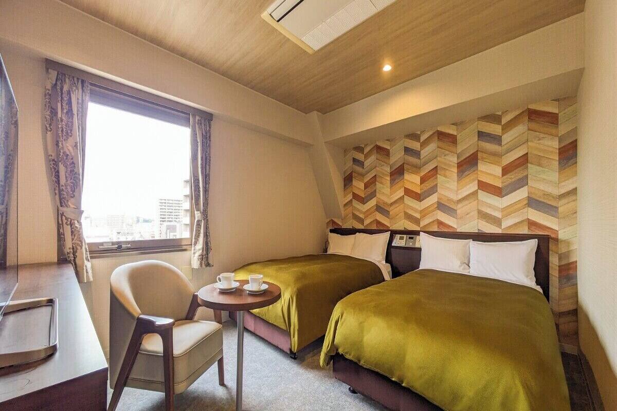 A hotel room with two beds, a desk, a chair, and a small table with cups.