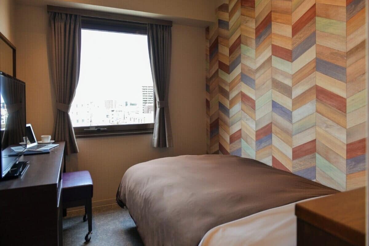 A hotel room with a bed, a desk with a laptop, a chair, and a window with curtains.