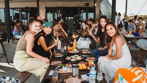 Un grupo de personas sentadas en un área de comedor al aire libre, con diversos alimentos y bebidas sobre la mesa.