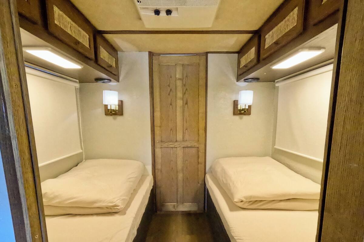 A narrow corridor with two beds on either side, wooden paneling on the walls, and wall-mounted lights.