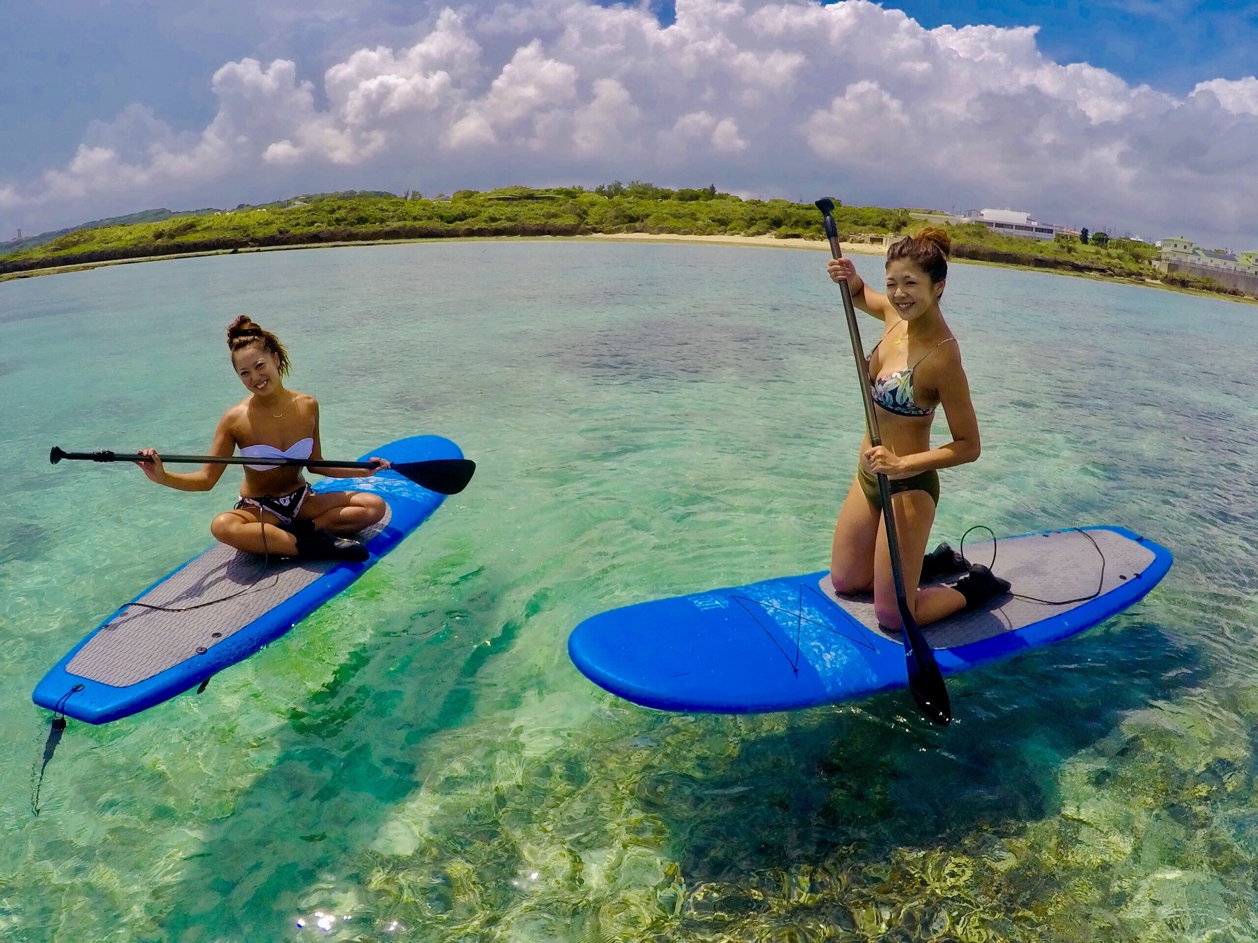 Two people paddleboarding on clear turquoise water with a beach and greenery in the background.