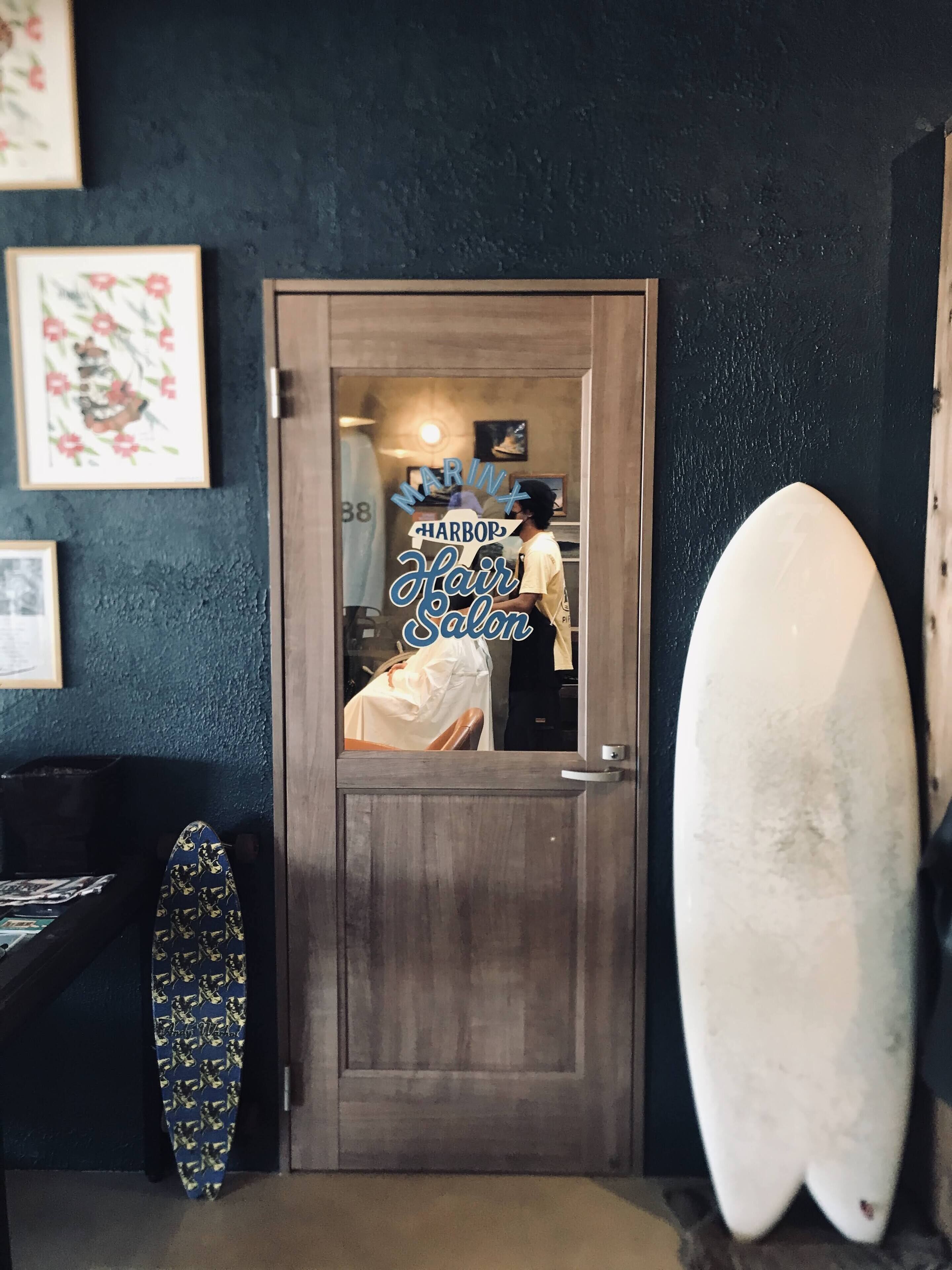 Eine Tür, in einem Spiegel darin sichtbar: ein Friseursalon, ein Surfbrett und ein gemustertes Handtuch.