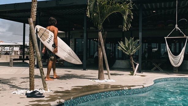 Un hombre lleva una tabla de surf cerca de una piscina con palmeras y una zona cubierta al fondo.