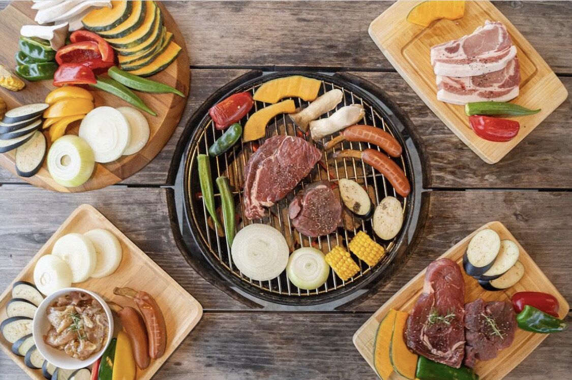 Une variété de viandes et de légumes grillés, avec d’autres plats disposés sur des planches en bois autour du grill.