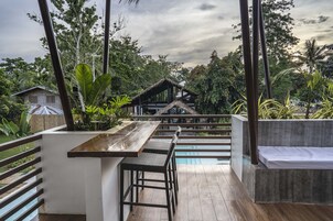 Una terraza de madera con área de bar, piscina y vistas a una casa tradicional de madera rodeada de abundante vegetación.
