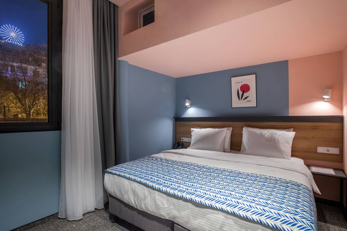 A hotel room with a bed, a nightstand, a wall-mounted lamp, and a window with curtains.