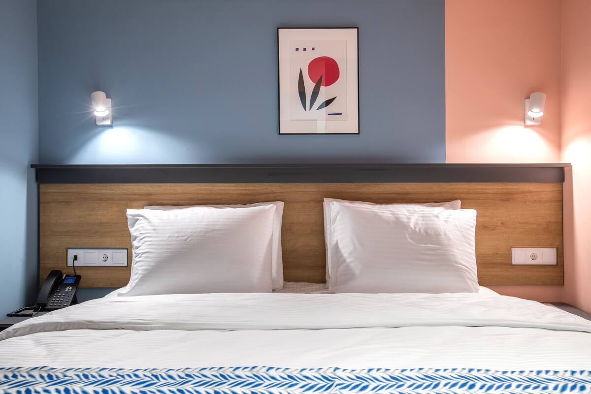 A hotel room with a wooden headboard, white bedding, a framed artwork on the wall, and a bedside phone.