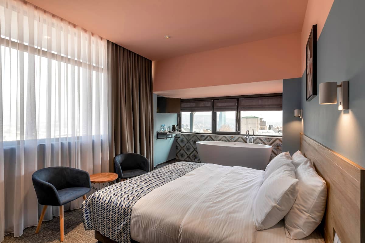 A hotel room with a bed, a chair, a small table, a bathtub, and a city view.