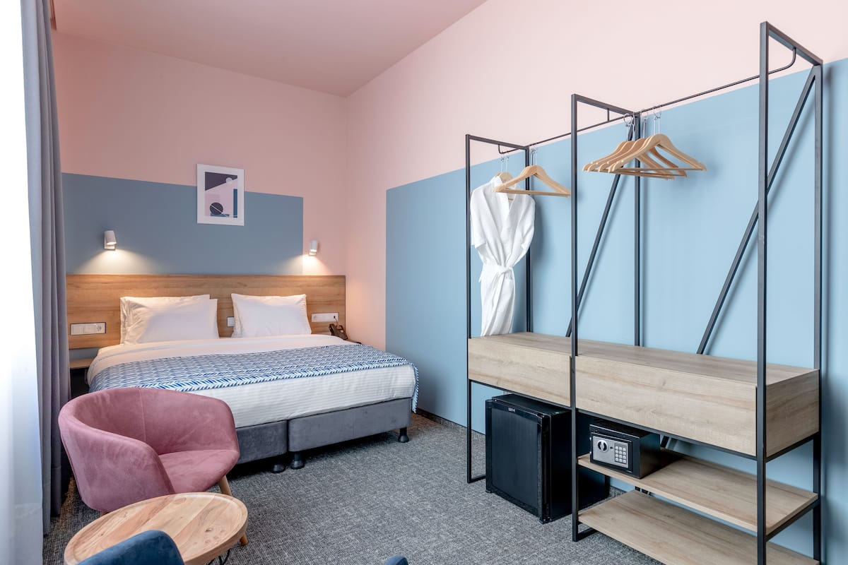 A hotel room with a bed, a chair, a small table, a wardrobe, and a desk with a computer.