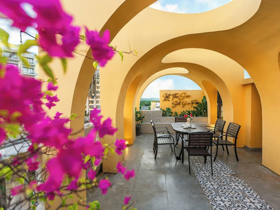 Un patio iluminado por el sol, con una mesa y sillas, rodeado de arcos y flores coloridas.