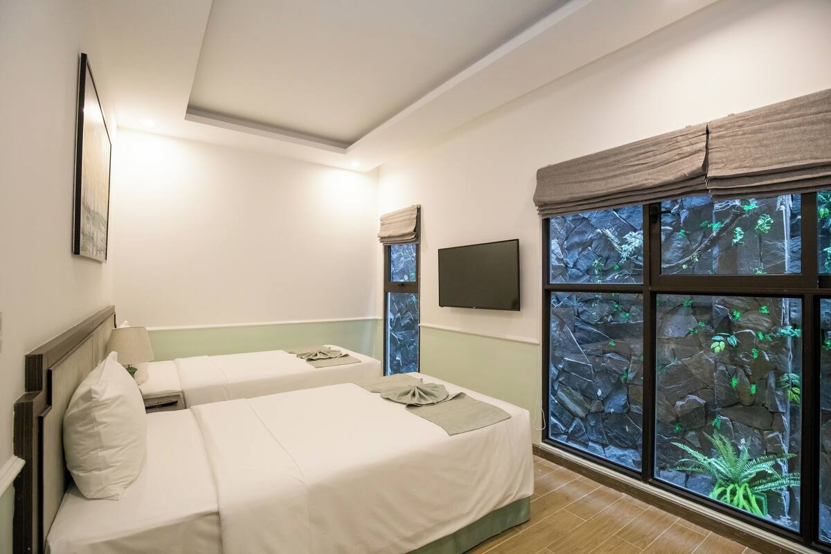 A hotel room with two beds, a mounted TV, and a large window with a view of rocks and greenery.