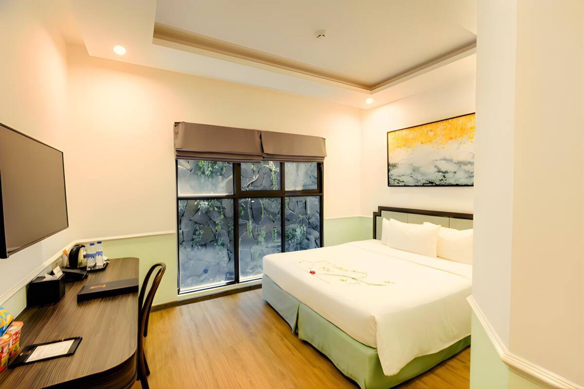 A hotel room with a bed, a desk with a chair, a TV, and a window with a view of trees.