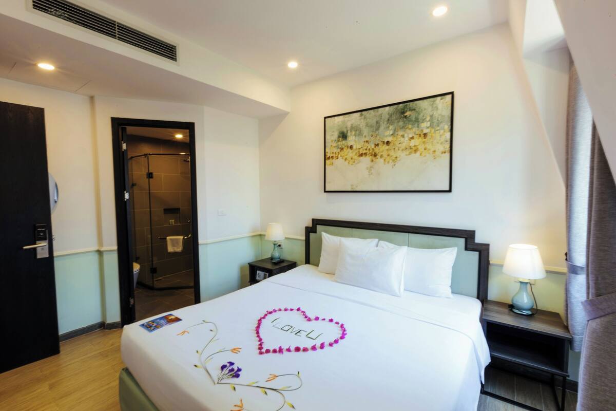 A hotel room with a bed, a nightstand with a lamp, a shower area, and a painting on the wall.