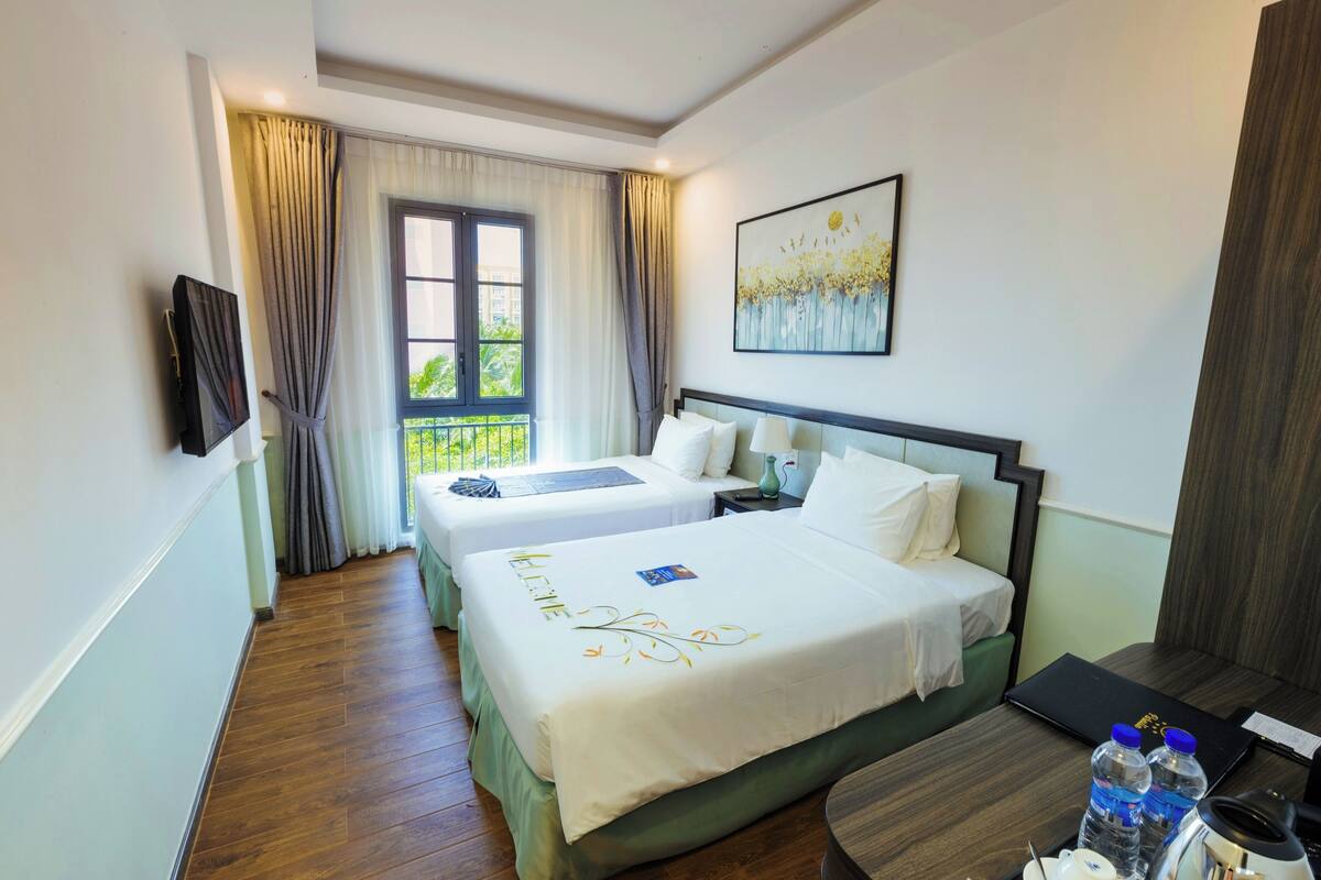 A hotel room with two beds, a desk, a chair, a TV, a window with curtains, and a painting on the wall.