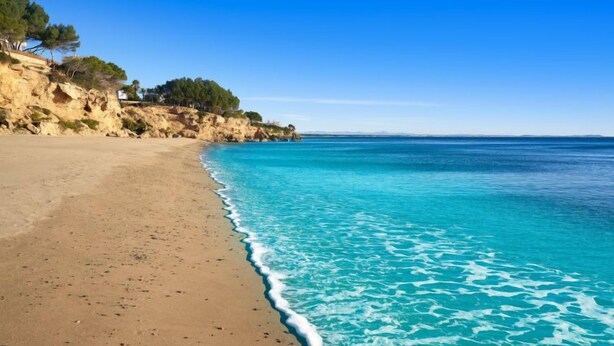 Una playa con agua turquesa cristalina, orilla de arena y acantilados rocosos con vegetación escasa.