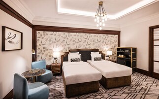 Hotelkamer met twee bedden, een blauwe fauteuil, een klein rond tafeltje, een kroonluchter en een decoratief wandpaneel.