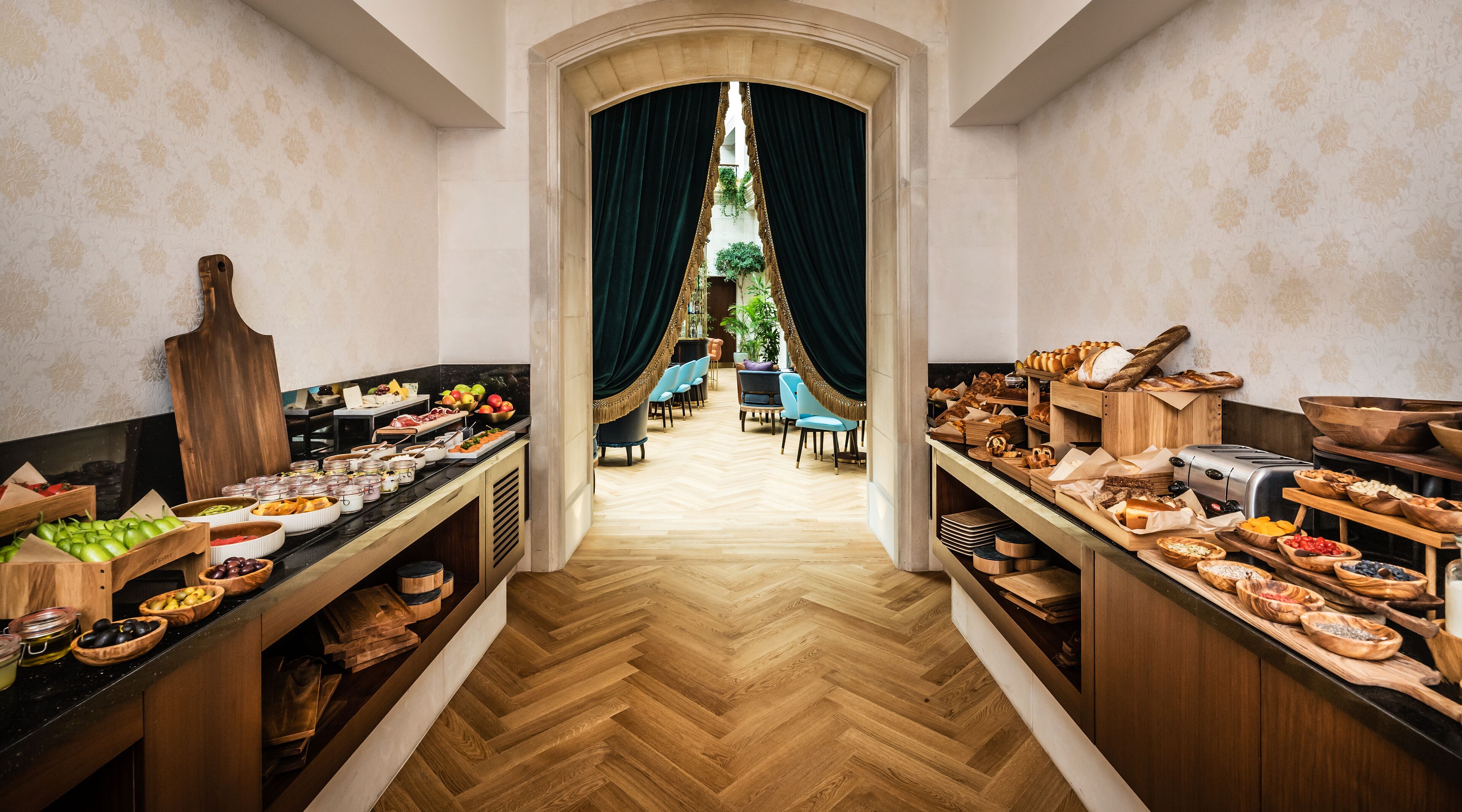 A hotel dining area with a long wooden herringbone floor, a central archway, and a view into a dining room with blue chairs and a plant.