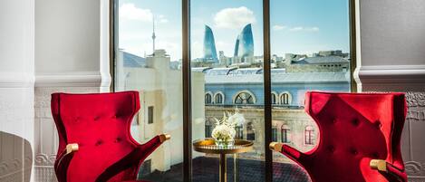 A room with two red velvet armchairs, a small round table with a vase of flowers, and a large arched window offering a view of a cityscape.