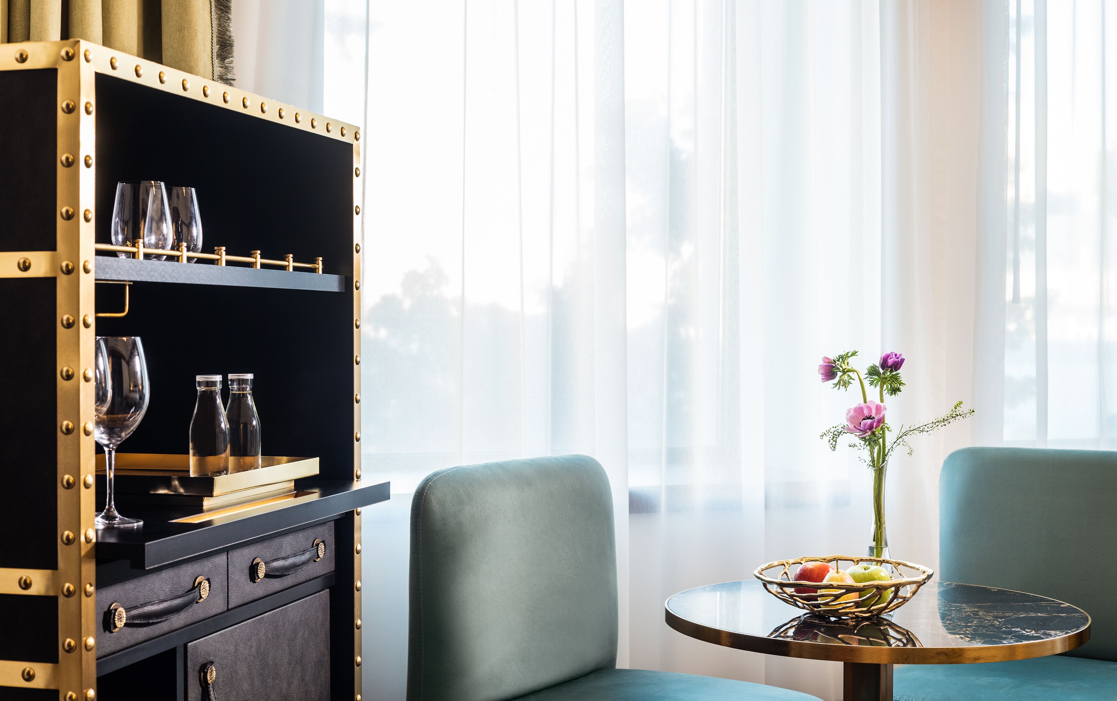Een kamer met een zwart metalen kast met glazen, een tafel met een fruitschaal, en twee groene bekleden stoelen.