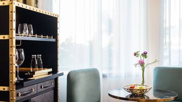 Een kamer met een zwart metalen kast met glazen, een tafel met een fruitschaal, en twee groene bekleden stoelen.