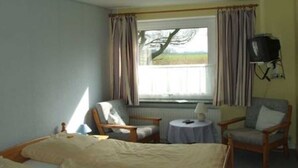 Un dormitorio con una cama, dos sillones, una mesita, una ventana con cortinas y un televisor.