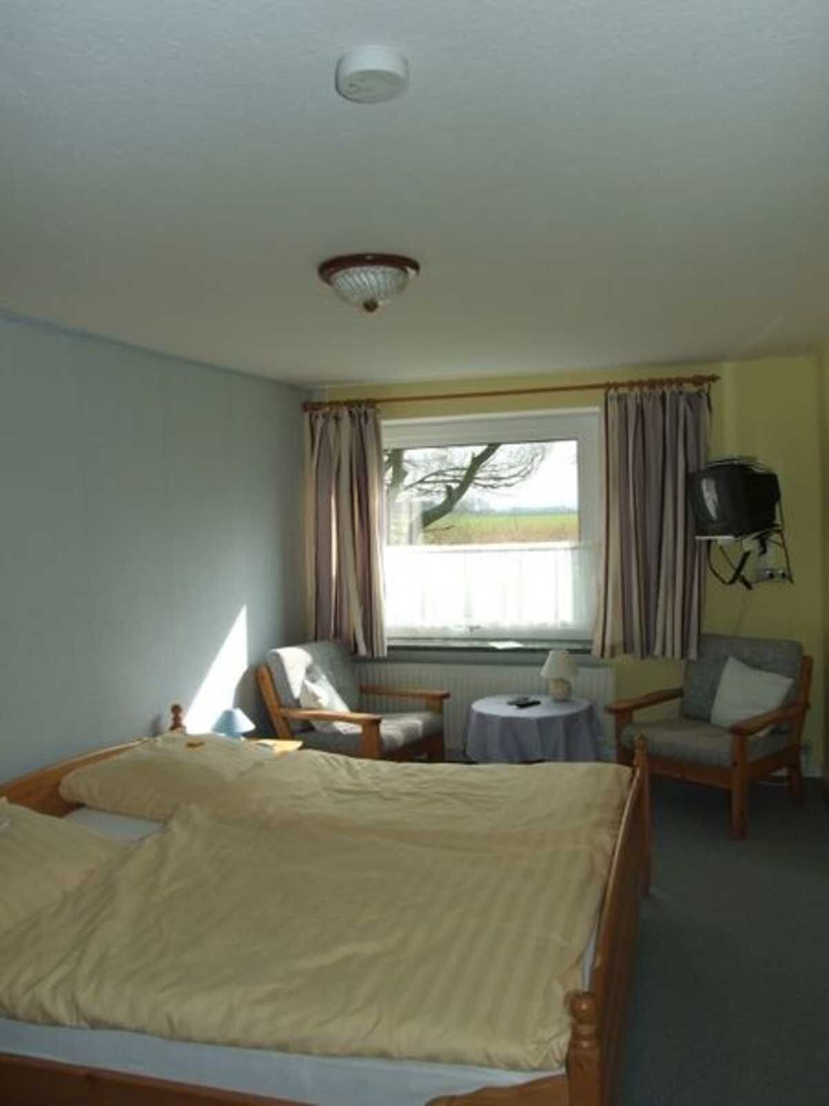 Une chambre à coucher comprenant un lit, deux fauteuils, une petite table, une fenêtre avec des rideaux et un téléviseur.