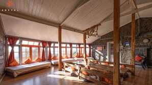 Una habitación amplia con bancos de madera y camas, ventanas grandes con cortinas naranjas y una pared de piedra.